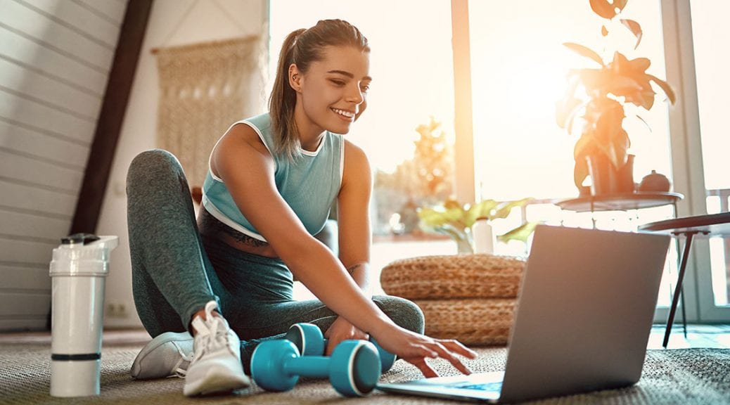 A sporty woman in sportswear is sitting on the floor with dumbbells and a protein shake or a bottle of water and is using a laptop at home in the living room. Sport and recreation concept; blog: 11 Low-Impact Exercises to Try